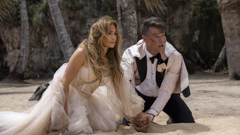 "Shotgun Wedding - Ein knallhartes Team": Darcy (Jennifer Lopez) und Tom (Josh Duhamel) knien in ramponierter Hochzeitskleidung am Strand nebeneinander und halten sich gegenseitig an der Hand. Beide schauen panisch in die gleiche Richtung. Im Hintergrund stehen Palmen.