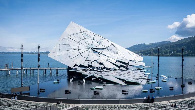 "Von der Seebühne Bregenz: La Traviata": Das Bühnenbild der Bregenzer Seefestspiele besteht aus einer großen Wasserfläche. Darauf befindet sich ein überdimensionierter Spiegel, der in viele Einzelteile zerfällt. Im Vordergrund befinden sich die Sitzplätze für die Zuschauer, im Hintergrund liegt der See.