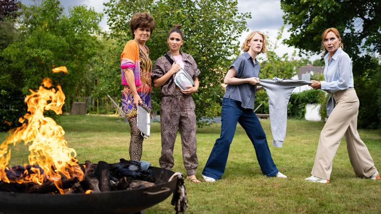 "Plötzlich Schwester": Im Garten von Sandra Giesinger: Gabi Koslowski (Anita Vulesica) hält ein silbernes Tablett in einer Hand und steht neben ihrer Tochter Sandy (Cosima Henman), die mit beiden Händen ihren Brustbeutel festhält. Neben ihnen ziehen Sandra Giesinger (Klara Lange) und ihre Mutter Monika (Andrea Sawatzki) an einem grauen Pullover. Alle vier blicken verwirrt nach vorne, wo ein großes Lagerfeuer zu sehen ist.