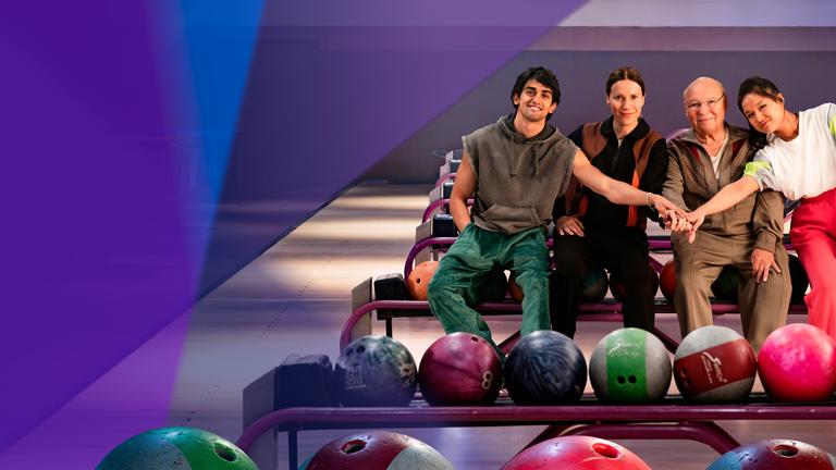 "Die Abräumer": Djamal (Mido Kotaini), Karo (Bettina Lamprecht), Freddy (Hans-Joachim Heist) und Shu (Jing Xiang) sitzen nebeneinander auf dem Ballrücklauf-Gestell der Bowlingbahn. Sie halten ihre Hände zusammen und sehen lächelnd in die Kamera.