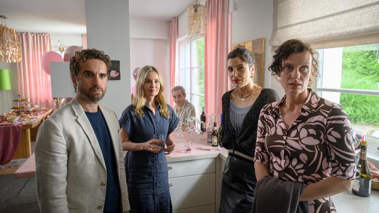 "Die Bachmanns - Gefahr im Verzug": Miriam (Stefanie Stappenbeck) steht auf Galinas Geburtstagsparty zusammen mit Markus (Oliver Wnuk), Andrea (Sophia Mercedes Burtscher) und Lydia (Effi Rabsilber) in der Küche, Juan (Oscar Ortega Sánchez) sitzt im Hintergrund am Esstisch. Alle schauen in die selbe Richtung, seitlich nach vorne.