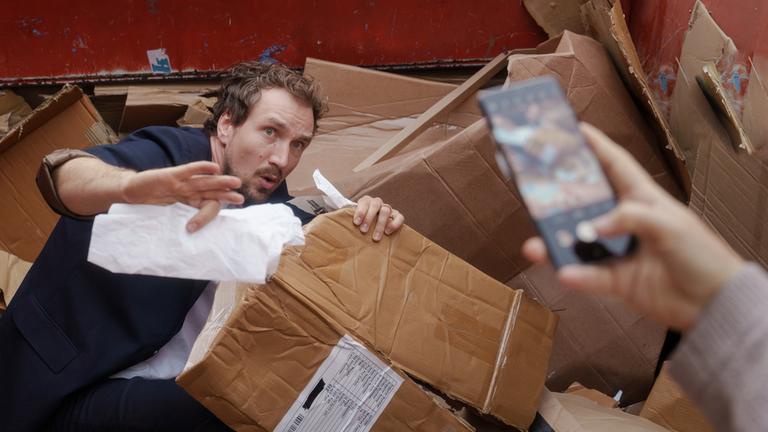 "Fang mich doch!": Georg Sommer (Jan Krauter) hockt im Altpapiercontainer. Er blickt erschrocken seitlich nach oben und macht eine abwehrende Geste. Genau dort hält eine Hand ein Smartphone und filmt ihn.