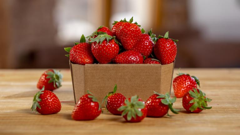 "Erdbeeren – Genuss mit Beigeschmack": Auf einem Holztisch steht eine Schale aus Pappe mit frischen Erdbeeren. Mehrere Früchte liegen davor, während der Hintergrund weich verschwimmt.