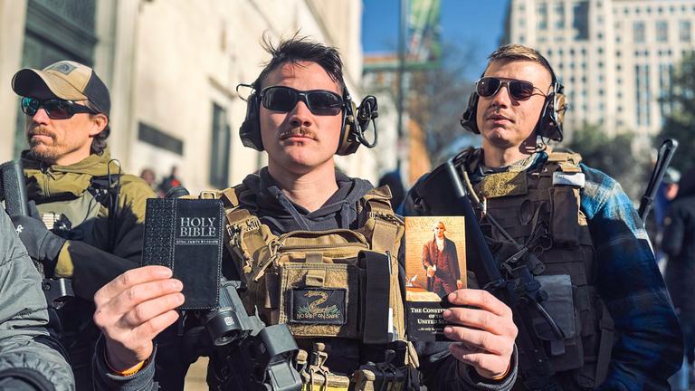  "Amerika - Traum und Wirklichkeit - Spaltung": Drei Männer mittleren Alters mit Sonnenbrille laufen in der Stadt. Alle sind mit Schutzwesten und Kopfhörern ausgestattet und bewaffnet. Der Mann in der Mitte hält die Bibel und die amerikanische Verfassung in die Kamera.