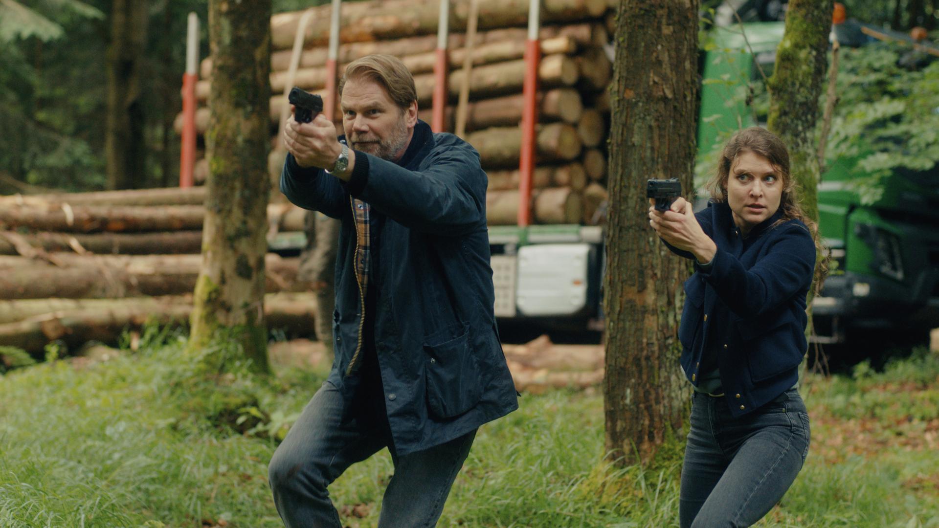  "Erzgebirgskrimi - Mordholz": Im Wald stehen die Ermittler Robert Winkler (Kai Scheve) und Karina Szabo (Lara Mandoki) Seite an Seite mit gezückten Waffen. Ihr Blick richtet sich konzentriert in die gleiche Richtung. Im Hintergrund türmen sich Baumstämme auf einem Lastwagen.