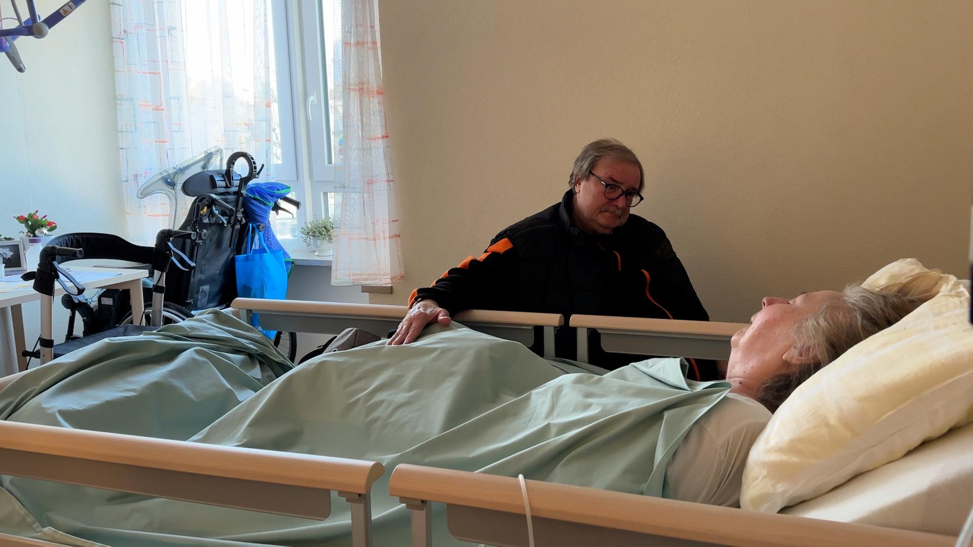 "37°: Koma: zwischen Leben und Tod": Ein älterer Mann sitzt am Krankenhausbett seiner Frau und schaut sie mitfühlend an. Seine Hand liegt auf der Bettdecke und berührt ihr Bein
