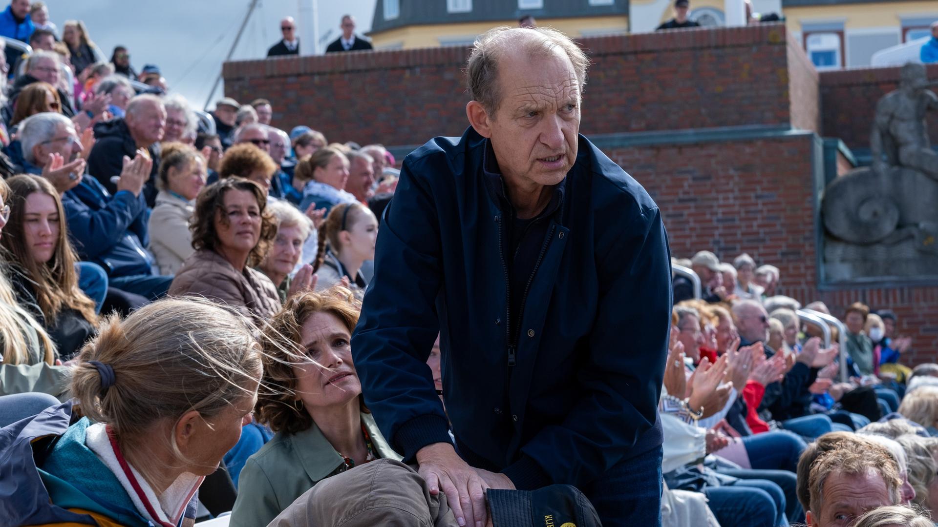 "Nord Nord Mord - Sievers und der letzte Tango": Im Freien auf der Zuschauerrängen: Carl Sievers (Peter Heinrich Brix) steht neben einem nach vorne gebeugtem Mann und fühlt mit seiner Hand seinen Puls am Hals. Tabea sitzt hinter ihnen und schaut angespannt zu Sievers. Rundherum sitzen Zuschauer, die zu Sievers und dem Toten schauen.
