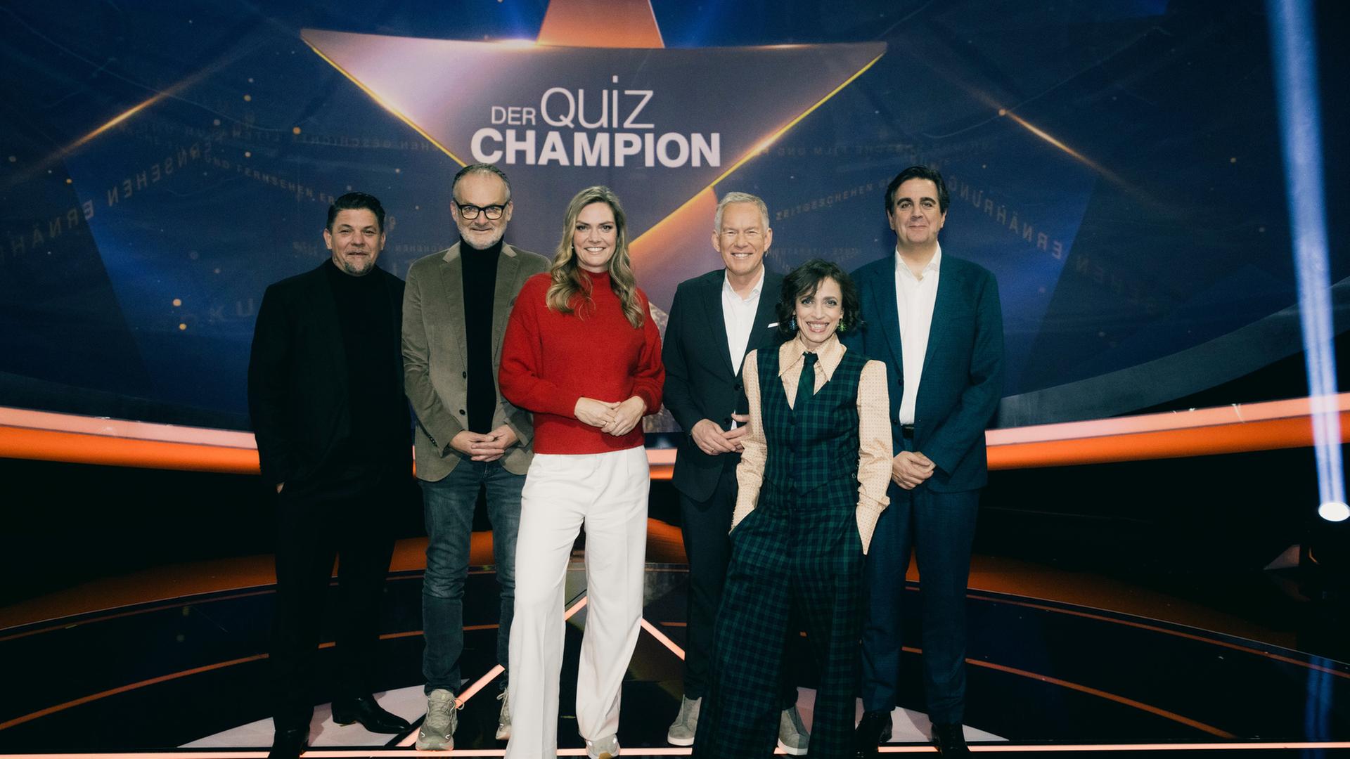"Der Quiz-Champion": Gruppenfoto: Tim Mälzer, Frank Plasberg, Julia Scharf, Johannes B. Kerner, Anna Thalbach, Bastian Pastewka stehen vor dem Sendungslogo im Studio und lächeln in die Kamera.