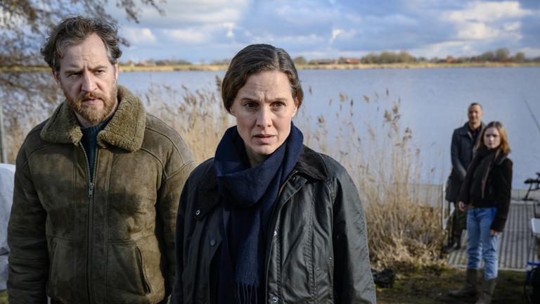 "Ostfriesensturm": Am Ufer eines Sees sieht Frank Weller (Tom Radisch) zu Ann Kathrin Klaasen (Picco von Groote), die neben ihm steht und nachdenklich schaut. Im Hintergrund stehen Rupert (Barnaby Metschurat) und Kollegin Jessi Jaminski (Sofie Eifertinger) beim Steg und schauen abwartend in Ann Kathrins Richtung.
