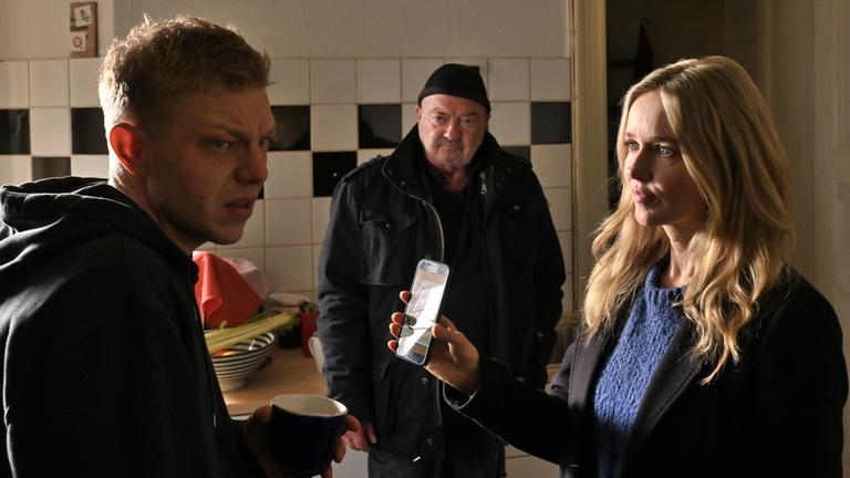 "Ein starkes Team - Mörderische Gier": In einer Küche: Linett Wachow (Stefanie Stappenbeck) zeigt Falk Becker (Leonard Kunz) ein Foto auf ihrem Mobiltelefon. Otto Garber (Florian Martens) blickt zu den beiden rüber.