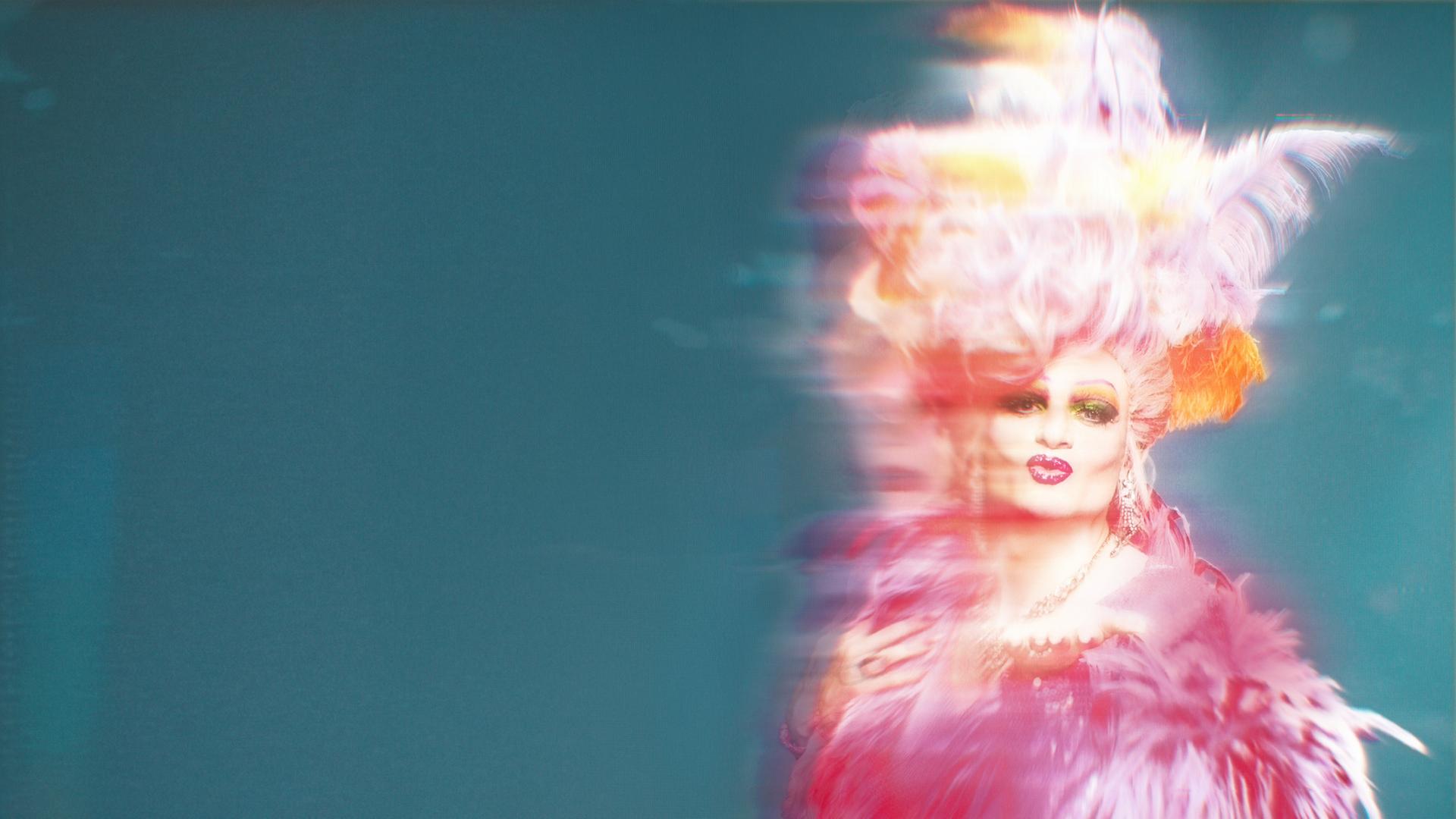 "Olivia": Olivia Jones/Oliver Knöbel (Johannes Hegemann) trägt ein auffälliges, pink‑ und lilafarbenes Federkostüm mit opulentem Kopfschmuck und steht mit Blick in die Kamera vor einem türkisfarbenen Hintergrund.