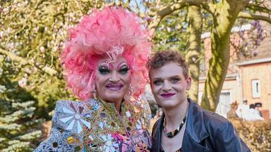 "Olivia": Olivia Jones/Oliver Knöbel (Johannes Hegemann) trägt ein auffälliges, pink‑ und lilafarbenes Federkostüm mit opulentem Kopfschmuck und steht mit Blick in die Kamera vor einem türkisfarbenen Hintergrund.