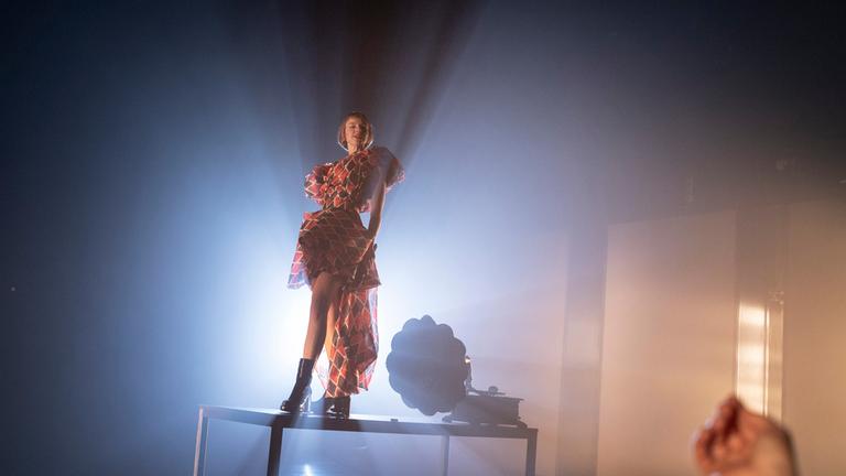 "Mephisto": Eine Frau steht auf einem metallenen Podest und ist stark von hinten beleuchtet, sodass Lichtstrahlen nach vorn ausfächern. Sie trägt ein asymmetrisches, mehrlagiges Kleid und Stiefel. Rechts ragt die Hand einer weiteren Person ins Bild.