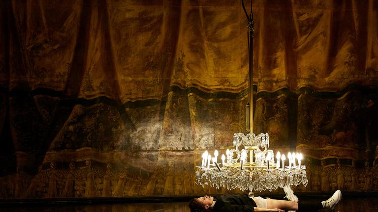 "Fräulein Else": Eine Frau liegt ausgestreckt auf dem Rücken auf einem hochglänzenden, schwarzen Bühnenboden. Direkt über ihrem Oberkörper hängt ein großer Kristalllüster mit vielen brennenden Kerzen, deren Licht sich auf der Bodenfläche spiegelt. Im Hintergrund hängt ein Bühnenvorhang in warmen Braun‑ und Goldtönen.