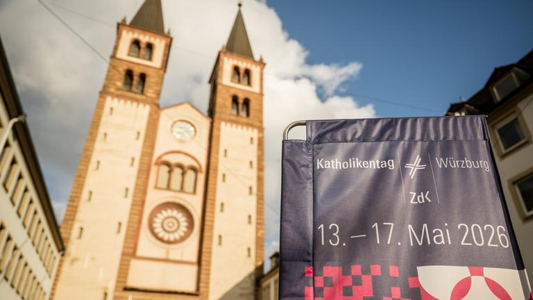 Ein Banner des Katholikentags hängt neben dem Würzburger Dom, am blauen Himmel ziehen weiße Wolken auf.