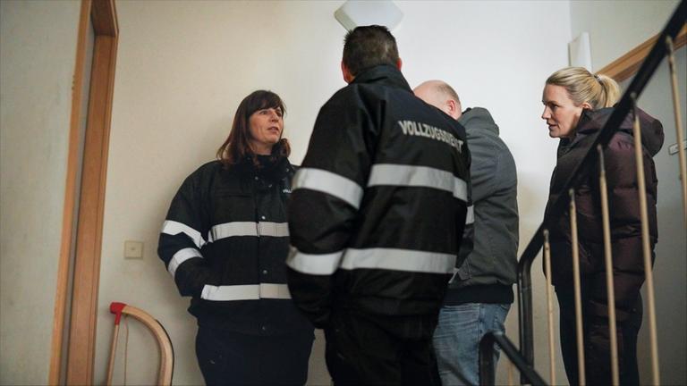 "Am Puls mit Sarah Tacke": Sarah steht mit drei Personen in einem Treppenhaus. Zwei Personen tragen Jacken mit der Aufschrift "Vollzugsbeamter".