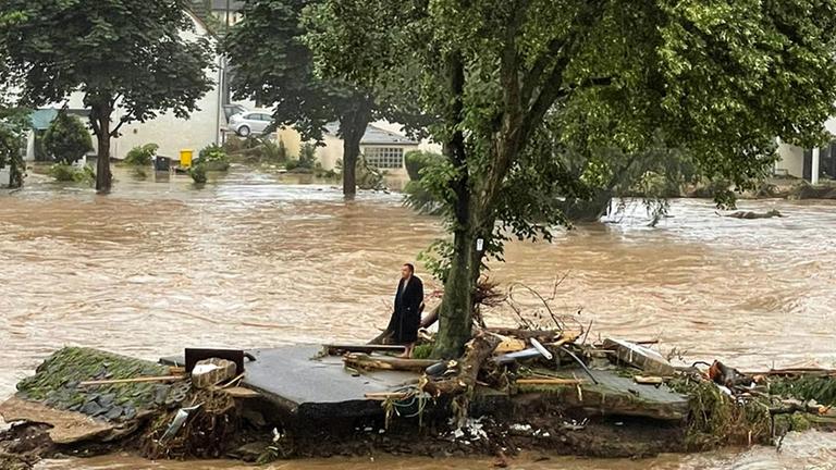 "TerraX History: Ein Tag im Juli - Ahrtalflut 2021": Ein Mann im Bademantel steht verloren auf einem Reststück Straße neben einem Baum. Um ihn herum nur braunes Wasser. Im Hintergrund stehen Häuser im Wasser.