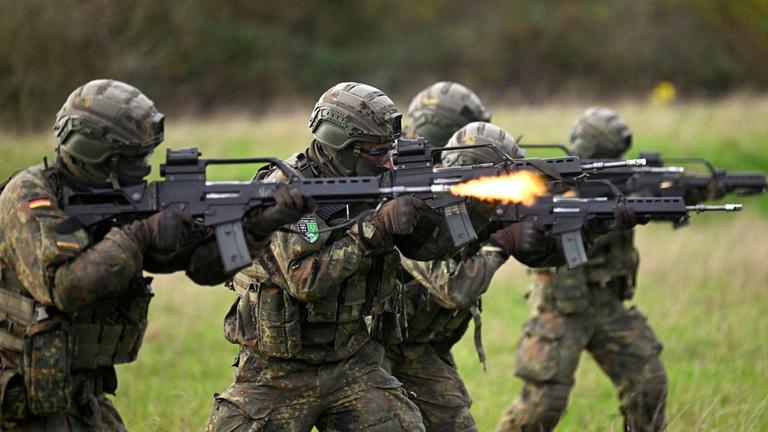  "Kriegstüchtig? 70 Jahre Bundeswehr": Fünf Soldaten der Bundeswehr stehen auf einer Wiese und feuern mit Gewehren.