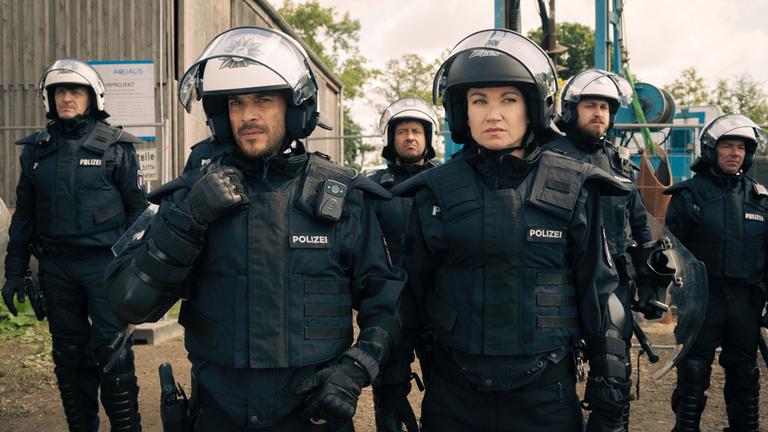 "Sarah Kohr – Bis auf den Grund": Polizeizugführer Till Ruprecht (Kostja Ullmann) und Sarah Kohr (Lisa Maria Potthoff) stehen in Montur der Bereitschaftspolizei nebeneinander vor einer Baustelle. Till Ruprecht greift an sein Funkgerät. Im Hintergrund befinden sich vier weitere Beamte (Komparsen).