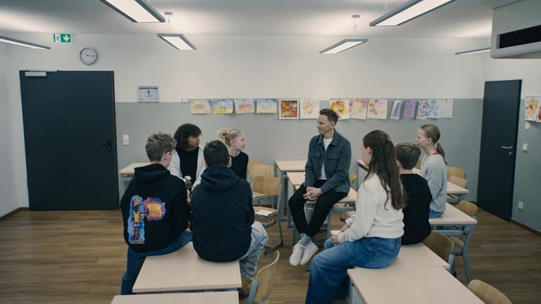"Die Wahrheit über Social Media - mit Jochen Breyer": Jochen Breyer sitzt in einem Klassenzimmer auf einem Tisch. Um ihn herum sitzen sieben Schüler und Schülerinnen ebenfalls auf Tischen.