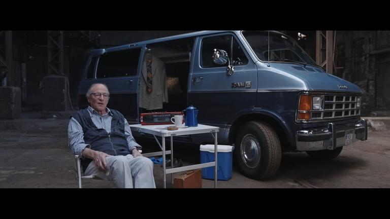 "Meine D-Mark: Als die DDR das Westgeld bekam": Hermann von Schrötter sitzt auf einem Campingstuhl vor einem blauen Kleinbus in einer Industriehalle. Vor ihm stehen ein Klapptisch und verschiedene Alltagsgegenstände.