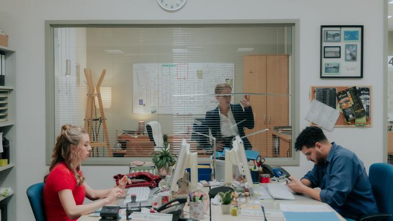 "Nine to Five - Das Amt - Ferienanlage Döberitz": Melissa (Anna Herrmann) und Orkan (Yasin El Harrouk) sitzen sich an ihren jeweiligen Schreibtischen gegenüber und blicken auf Bildschirme. Im Hintergrund schaut Ronja (Friederike Kempter) durch ihr Bürofenster auf die beiden.