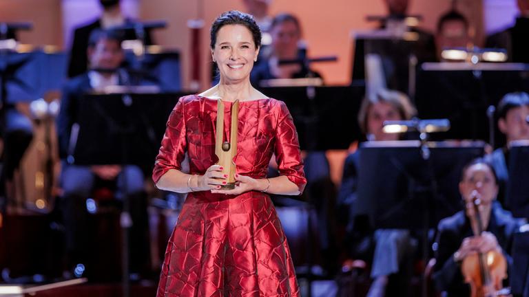 "OPUS KLASSIK 2026": Désirée Nosbusch steht vor einem Orchester und lächelt. In den Händen hält sie die OPUS KLASSIK-Trophäe.