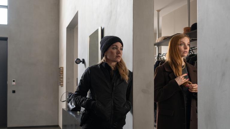 Sarah Kohr (Lisa Maria Potthoff), schwarz gekleidet, mit schwarzer Wollmütze und offenen Haaren, lehnt lässig, die Hände in den Jackentaschen, gegen eine Wand in einem modernen Haus. Auf der anderen Seite der Wand steht Mira Röver (Pheline Roggan) in einem braunen Mantel. Sie schaut angespannt.