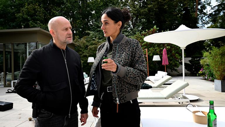 "Jenseits der Spree - Kontrollverlust": Robert Heffler (Jürgen Vogel) steht auf der Terrasse eines modernen Anwesens neben dem Pool und blickt zu Mavi Neumann (Aybi Era). Mavi Neumann hält eine grüne Flasche in der Hand und wendet sich Robert Heffler zu. Im Hintergrund stehen Sonnenschirme neben Liegestühlen.