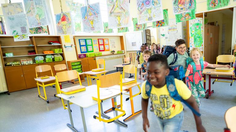 "37°: Unsere Klasse: Chaos und Chancen in der 1d": Schüler rennen mit Rucksäcken auf dem Rücken lächelnd in einer Reihe zwischen den Tischen durch in ein Klassenzimmer hinein.