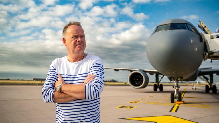 "Am Puls mit Christian Sievers: Welt in Dauerkrise – Was gibt uns Sicherheit?": Christian Sievers steht mit verschränkten Armen auf einem Flugfeld. Im Hintergrund befindet sich die Front eines Passagierflugzeugs.
