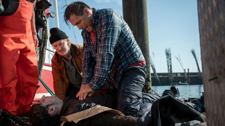 "Nord Nord Mord - Sievers und der Gesang der Wale": Am Hafen: Götz Fischer (Max Hopp) und Niels Schneider (Leo Meier) knien neben Hark "Harke" Petersen (Eren Yilmaz) und versuchen ihn zu reanimieren. Harke ist ganz blass und atmet nicht.