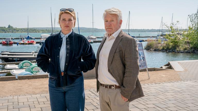 "Neues Land": Ina Sedlitz (Jördis Triebel) und Walter Sedlitz  (Jörg Schüttauf) stehen nebeneinander an einem gepflasterten Uferweg. Hinter ihnen liegen Boote auf einem See mit bewaldetem Ufer. Beide blicken ernst.