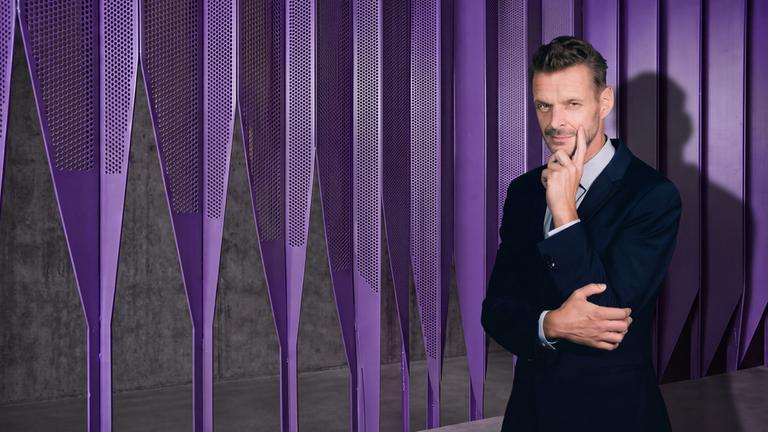 "JOURNAL DELUXE": Florian Schroeder steht auf der rechten Seite des des Bildes. Er hält seinen linken Zeigefinger an seine linke Wange uns stützt mit der rechten Hand den seinen linken Ellenbogen. Der Hintergrund ist lila.