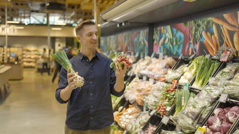 "plan b: next BITE: Gutes Essen geht auch günstig": Eric Mayer steht in der Gemüseabteilung im Supermarkt. In einer Hand hält er Frühlingszwiebeln, in der anderen Radieschen.