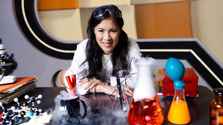 Mai Thi Nguyen-Kim sitzt im Studio von „MAITHINK X“ hinter einem Tisch mit verschiedenen Laborgefäßen und Versuchsgegenständen