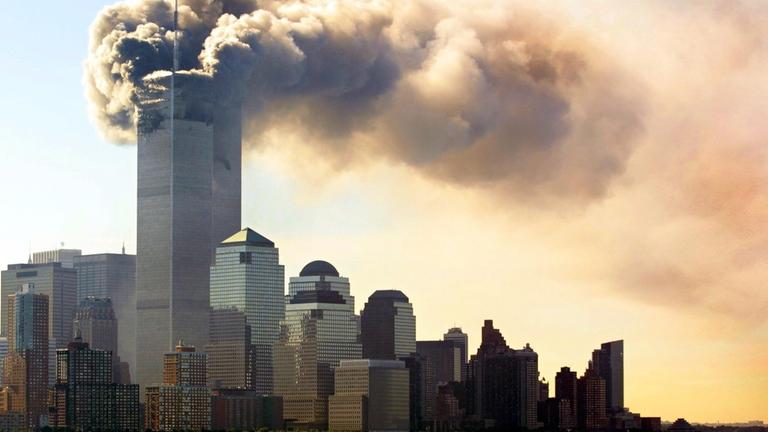 "9/11 - Der lange Schatten des Terrors - Spaltung": Panorama von Manhattan am 11. September 2001 mit Fokus auf das World Trade Center, dessen oberer Bereich von einer massiven Rauchwolke umhüllt ist. Die Rauchentwicklung dominiert die Skyline, während sich im Vordergrund Boote auf dem Wasser befinden.