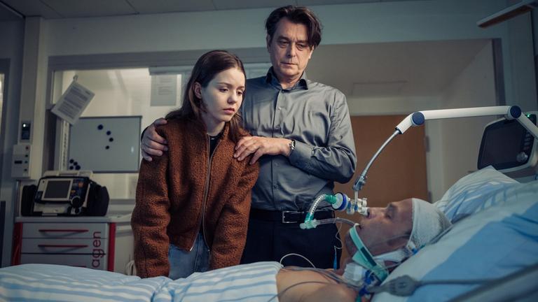 "Die Toten vom Bodensee - Im Schattenreich": Luna Oberländer (Fiona Katharina Neumeier) und Thomas Komlatschek (Hary Prinz) stehen bei Micha Oberländer (Matthias Koeberlin) am Krankenbett.