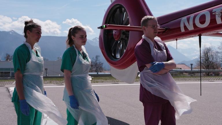 "Die Lebensretter von Murnau - Einsatz am Limit": Vor einem Bergpanorama bei Sonnenschein: Drei Pflegekräfte stehen neben dem Heckrotor eines Helikopters. Ihre OP-Schürzen wehen im Luftzug.