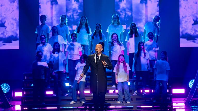 "Die Giovanni Zarrella Show – Kleine Stars ganz groß": Der Graf, Sänger der Band Unheilig, steht auf der Bühne und singt in ein Standmikrofon. Hinter ihm auf einer Showtreppe steht der Kinderchor Lucky Kids.