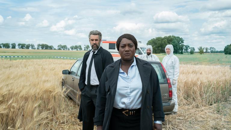  "Die Polizistin und die Todesstille": Boris Lambert (Tobias Oertel, l.) und Gloria Acheampong (Thelma Buabeng, 2.v.l.) stehen im Feld vor einem Wagen und schauen nach vorne. Neben dem Auto stehen zwei Spurensicherer (Komparsen) im Schutzanzügen, im Hintergrund parkt ein Rettungswagen.