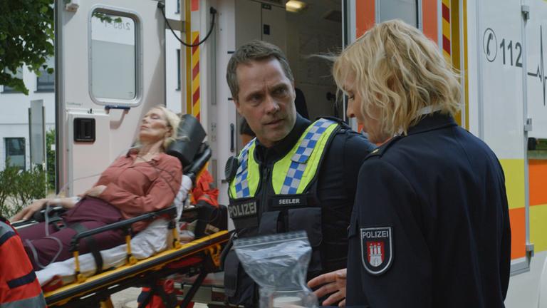"Notruf Hafenkante - The show must go on": Der Polizist Mattes Seeler (Matthias Schloo) und seine Kollegin Melanie Hansen (Sanna Englund) stehen seitlich hinter einem Rettungswagen. Melanie blickt auf den Beweisbeutel, den sie in der linken Hand hält. Mattes schaut sie an. Hinter den beiden liegt Karin Sievers (Katja Weitzenböck) auf der Krankentransportliege des RTW. Sie hat die Augen geschlossen.