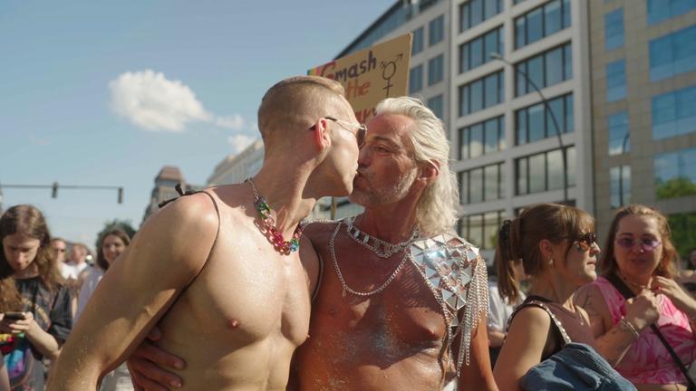 "Feindbild Queer - Kampf für die Vielfalt": Auf der Demonstration beim Christopher Steet Day  küssen sich zwei Männer. Um sie herum befinden sich weitere Personen, hinter ihnen steht ein mehrstöckiges Gebäude, die Sonne scheint.