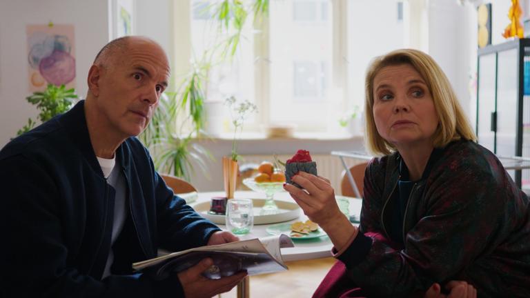 "Merz gegen Merz: Entscheidungen": Erik (Christoph Maria Herbst) und Anne (Annette Frier) sitzen gemeinsam am Esstisch ihres Sohnes. Während Erik eine Zeitschrift in den Händen hält, hat Anne ein kleines Kästchen in der Hand. Die beiden schauen neugierig zur Seite.