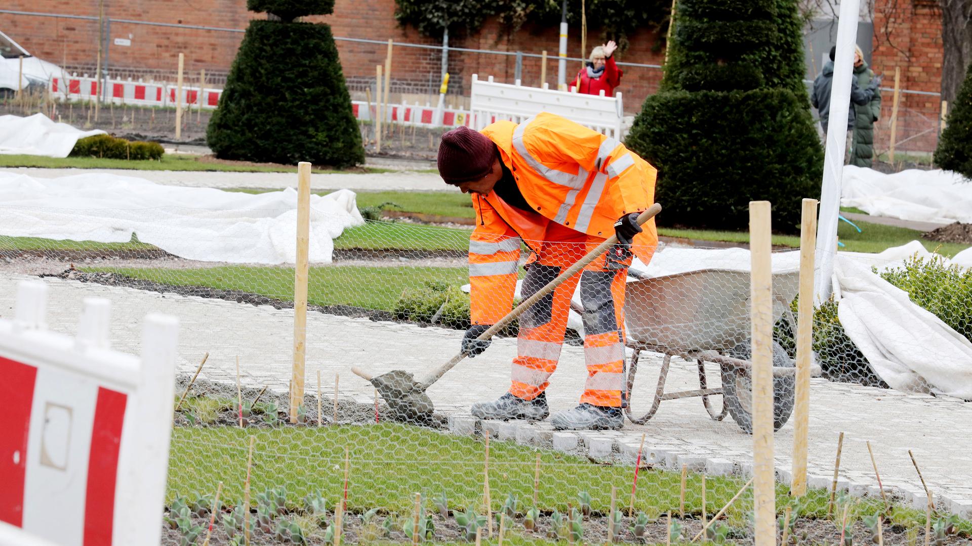 Ein Arbeiter in orangefarbener Arbeitskleidung mit Schaufel und Schubkarre befestigt einen Weg auf dem Gelände der Bundesgartenschau