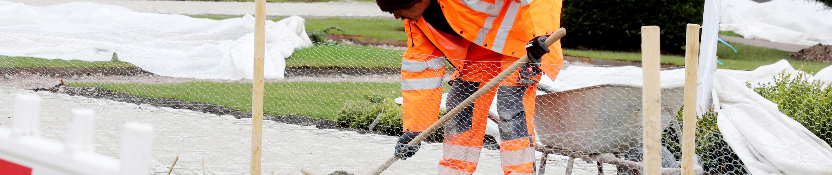 Ein Arbeiter in orangefarbener Arbeitskleidung mit Schaufel und Schubkarre befestigt einen Weg auf dem Gelände der Bundesgartenschau