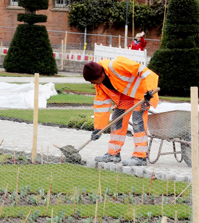 Ein Arbeiter in orangefarbener Arbeitskleidung mit Schaufel und Schubkarre befestigt einen Weg auf dem Gelände der Bundesgartenschau