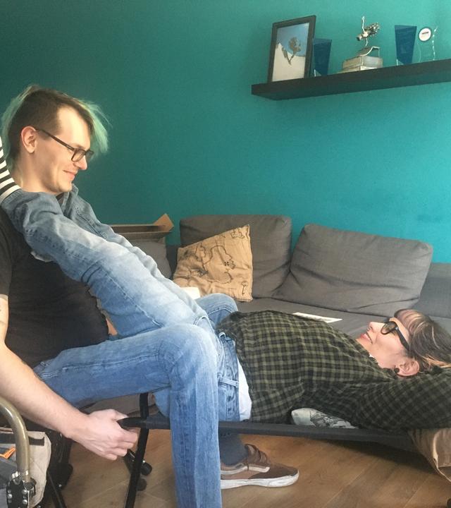 Ein Mann sitzt auf einem Stuhl. Seine Partnerin liegt auf einer Bank zwischen seinen Beinen. Ihre Füße liegen auf seinen Schultern.