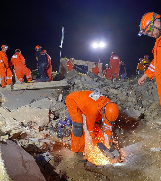 Männer in orangefarbenen Schutzanzügen suchen unter Trümmern nach Überlebenden.
