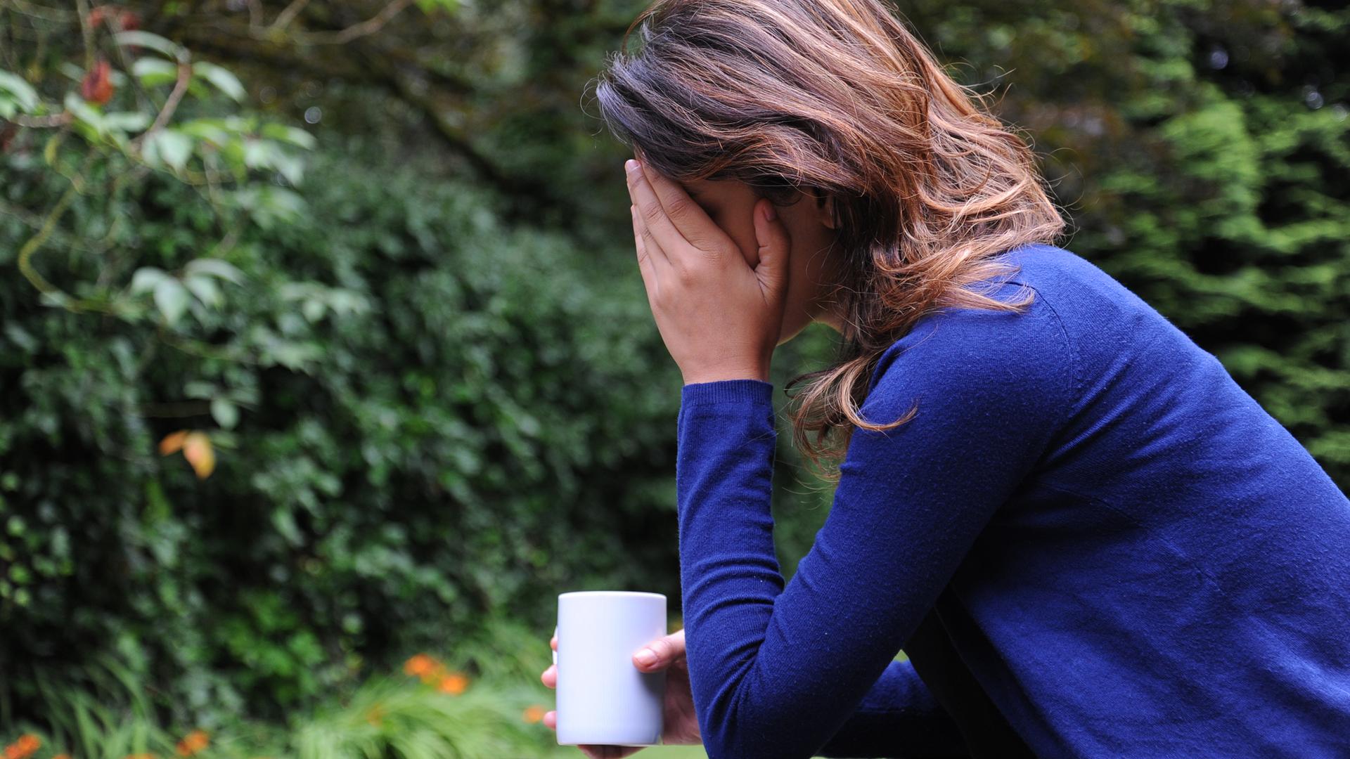 Frau sitzt mit Tasse draußen und hält sich mit einer Hand den kopf. Sie scheint deprimiert zu sein oder Schmerzen zu haben.